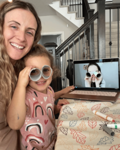 Preschooler and parent are participating in a virtual French preschooler class. The young girl is holding up a pair of DIY binoculars made as a fun craft during French class.