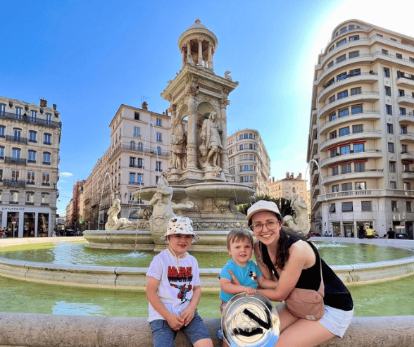 Madame Amy and her two young sons standing in front of a charming French fountain in Lyon.